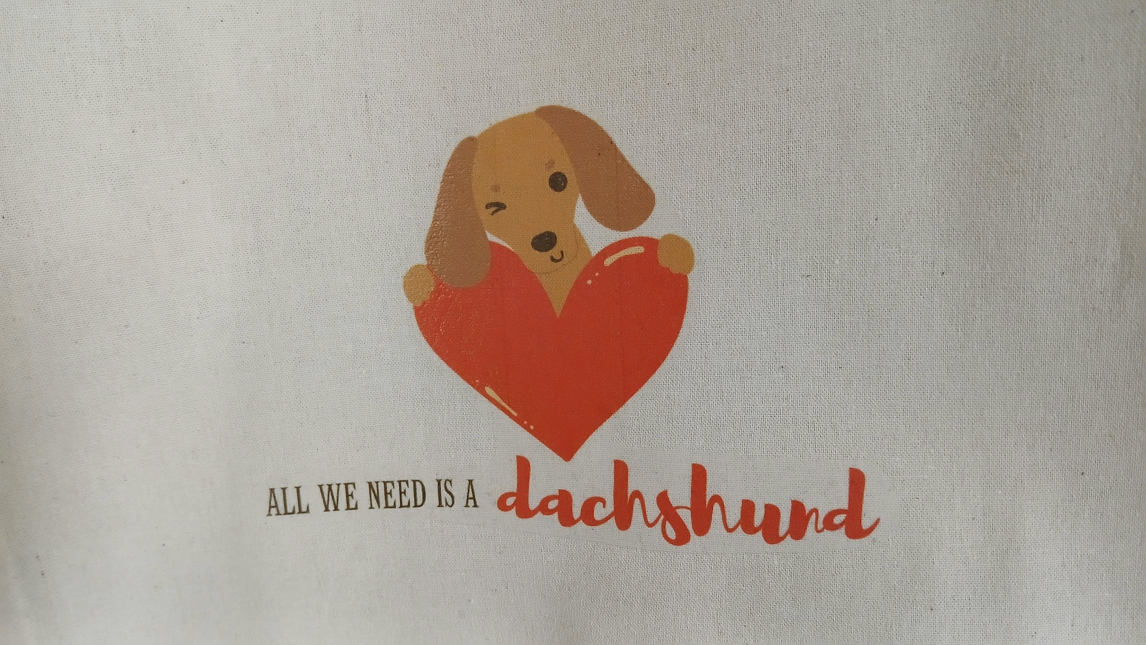 Tote bag with a dachshund holding a heart and text 'All we need is a dachshund' on a beige background.