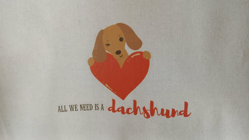 Tote bag with a dachshund holding a heart and text 'All we need is a dachshund' on a beige background.