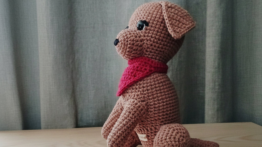 Crocheted Labrador plushie with a red scarf on a wooden surface with a curtain background